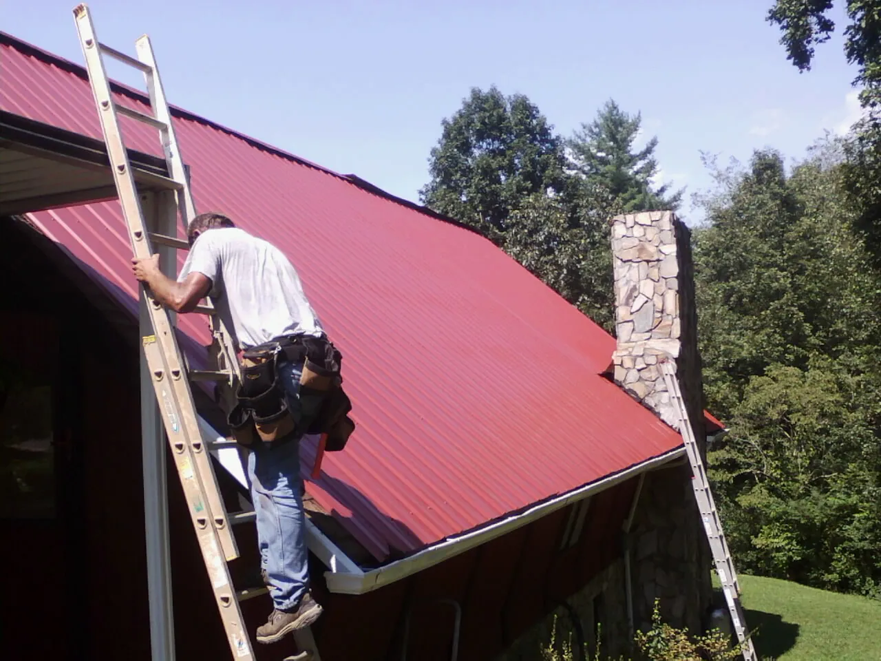 Roofer on a ladder beside a red metal roof and stone chimney — Soffit & Fascia Repair by Roofing Brothers in Eaton Center